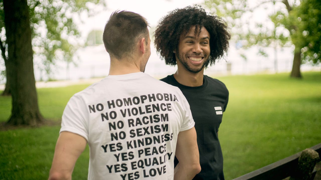 Two men smiling in a park, wearing T-shirts promoting equality and kindness.